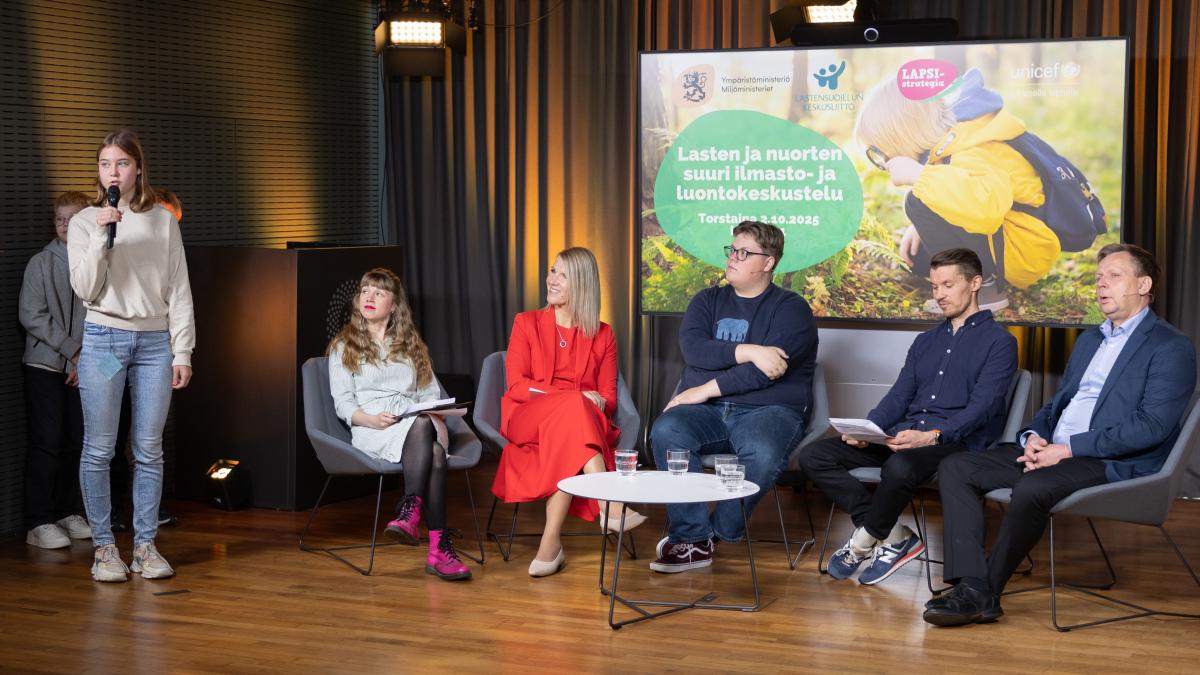 En elev i årskurs sex står med en mikrofon i handen och ställer en fråga till fem paneldeltagare som sitter i fåtöljer.