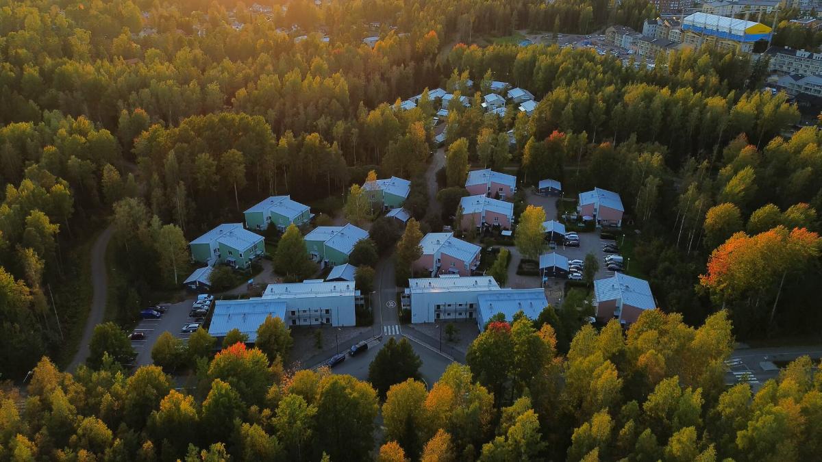 Flygbild av ett bostadsområde med grunda flervåningshus och småhus i en skogsmiljö.