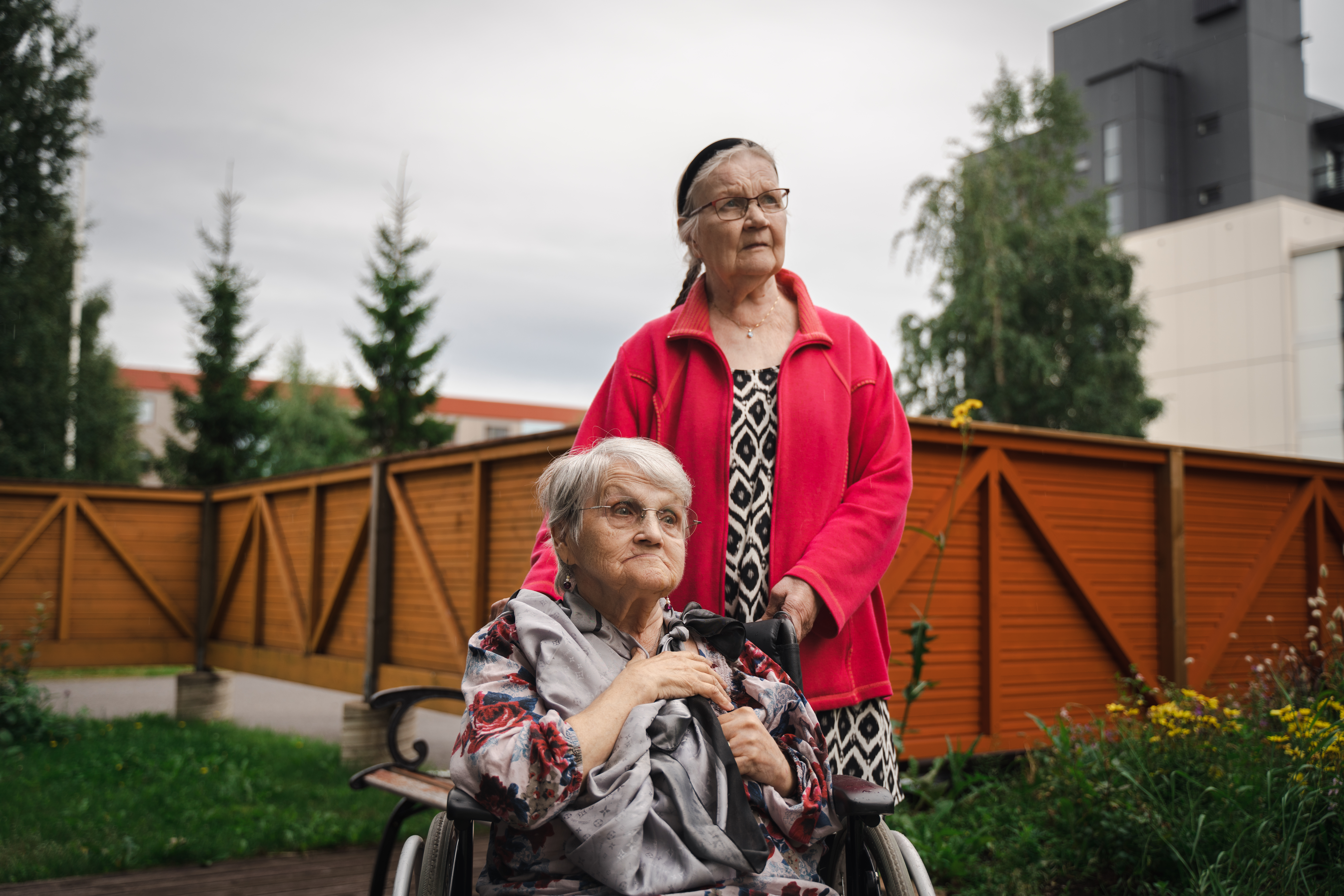 Kaksi iäkästä naista, joista toinen pyörätuolissa, istuvat puutarhassa. 