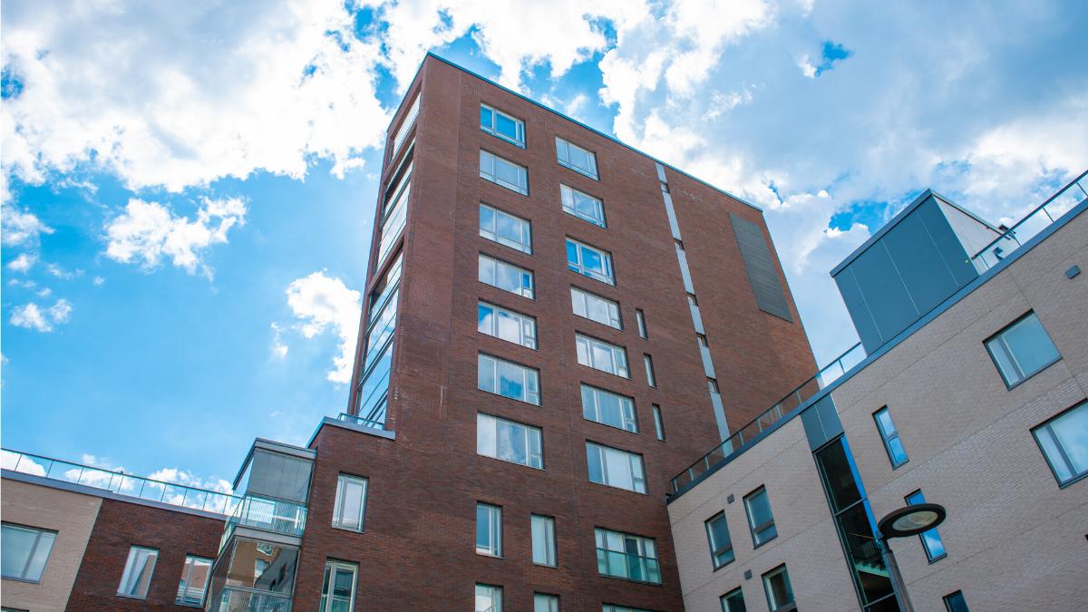 En röd tegelfastighet och en ljus höghusbyggnad mot en klar himmel. Byggnaden har stora fönster och balkonger. Bilden är tagen snett nerifrån och upp.