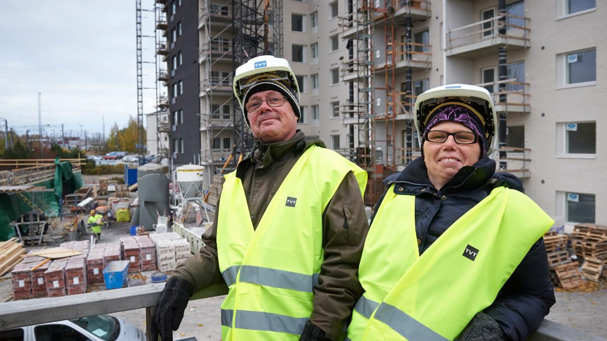 Två personer på en byggarbetsplats med gula skyddsvästar och hjälmar, i bakgrunden byggnation av flerbostadshus och byggmaterial.